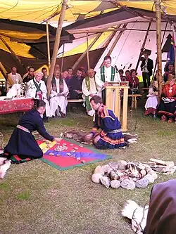 A church service held inside a large tent, with clergy seated behind a speaker at a lectern and two people in traditional Sámi clothing performing a ritual on the ground.