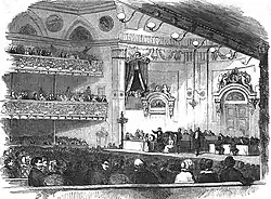 The interior of an elegant concert hall, an elegant audience, and on the stage, many men participating in the ceremony.