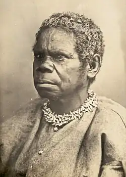 Portrait photograph of an Aboriginal woman, shown from the chest up, wearing a simple garment and a shell necklace, with short curly hair, facing to the left against a plain background.