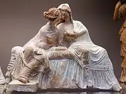 Terracotta statue of two women, wearing long dresses, seated and leaning on each other.