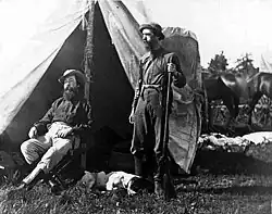 Black and white photograph of Dole and Nicolay in front of an open tent. Dole is seated, while Nicolay is standing, carrying a rifle. A dog sleeps by his feet.