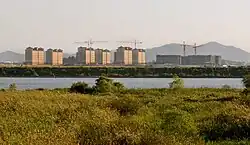A photograph of the Camp Humphreys military base in South Korea on the other side of a river, showing six buildings of seven-storey dorm barracks on the left, and shorter, wider buildings under construction on the right. Five cranes complete the skyline with a forested mountain range in the distance.