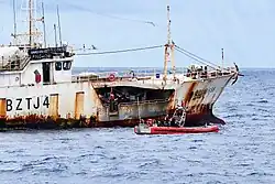 A rusty fishing ship with a small rubber boat pulled up alongside