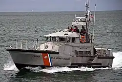 A grey metal boat moves through water toward the left. It features a diagonal red, white, and blue stripe, the text 'U.S. COAST GUARD', and the number '47289' on its hull. Crew members in life jackets are visible on the upper open bridge.