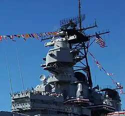 A large tower on a ship, on top is a radar antenna with an enclosed lookout space below the radar and a ship bridge below the enclosed space. Several people can be seen standing on the top of the bridge.