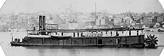Black-and-white photograph of a 19th-century steamboat in a river. The far riverbank in the background is lined with buildings covering a sloping bluff.
