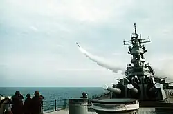 A forward view of a ship with large gun barrels pointing forward. To the left, a missile is flying away from the ship