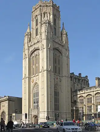 A tall stone nineteenth-century building with shields on the visible sides and a pepperpot upper storey. In front, traffic and pedestrians on a busy street.