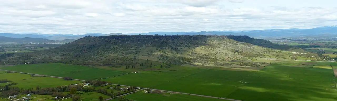 A large flat topped mesa with trees scattered on its flanks and farmland below it