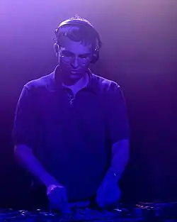 Vegyn performing on stage at a DJ set in Wigwam bar, Dublin, Ireland. He is lit entirely by blue stage lighting, and standing over a table with a CDJ on it.