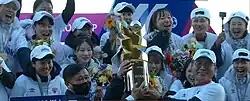 Female football players wearing white T-shirts and white baseball caps smile and laugh as they lift a golden trophy in the shape of the letters W and K