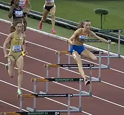 Photo of an athletics track with hurdles seen from an angle with Femke Bol jumping over a hurdle, followed by Yanique Haye-Smith, Eileen Demes, and two other athletes whose faces are not in the pictre