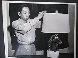 Image depicts a black and white photograph of Wah Chang adjusting a detail on a lampshade sculpture