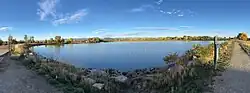This stitched panorama looks west from the east shore path at Waneka Lake in Lafayette, Colorado, with riprap and grasses in the foreground, calm reservoir waters at center, a loop trail and signpost at right, and cottonwoods in fall color backed by the Front Range under a clear evening sky.