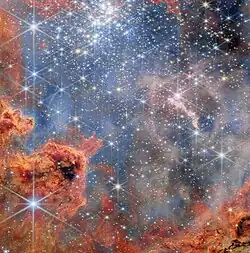 A cluster of stars inside a large nebula. The clouds of gas and dust are predominantly bright red in colour and wispy, akin to flames. They are clumped in the bottom-left corner. Other clouds, deeper in the cluster behind many of the stars, appear pale pink. The stars are concentrated in the top half of the image and are mostly small, bright white and six-pointed. They cast blue light over the nebula. Other stars with very long spikes surrounding them lie in the foreground.