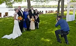 A man in a blue shirt and dark pants at right crouches as he takes a picture of a group of people at left in formal wear, with a woman in a white wedding dress at the rightmost. Behind them are tall trees and a large lake