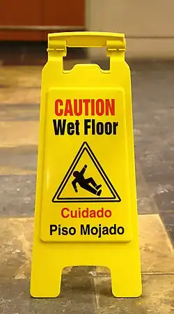 Sign reading "CAUTION Wet Floor" at the top, with a translation of the warning to Spanish at the bottom, and a pictogram in the center depicting a person slipping and falling over