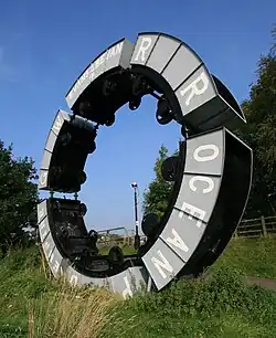 Modern sculpture using wheeled tubs from mineral mining attached to each other forming a raised circle against a bright blue sky