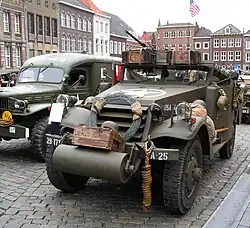 Photo of an M3 Scout Car from the front, outdoors on a city street with other military vehicles. It has a wide cylindrical object mounted on front beneath the radiator grill. A coil of barbed wire and coiled rope are also mounted.