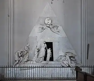 White marble pyramid-shaped tomb with sculpted figures mourning around entrance and medallion portrait at top
