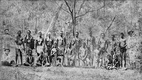 About 20 Aboriginal men with painted bodies holding spears pictured in two rows, with the back row standing and the front row sitting or kneeling. Three white men stand to the sides.