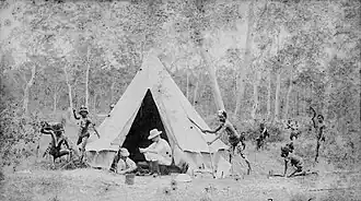 Two men sit in a white triangular tent as Indigenous men holding spears approach them