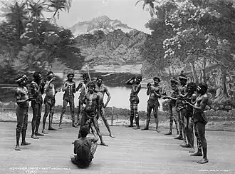 Photograph of a group of Aboriginal men with decorative painting on their torsos standing on a stage in front of a backdrop showing a rainforest and mountain