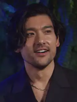 Will Sharpe wearing a dark shirt and a chain on his neck, grinning and looking left of camera