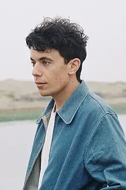 Side-profile photograph of a man stood by a lake dressed in a denim jacket facing to the left of the frame