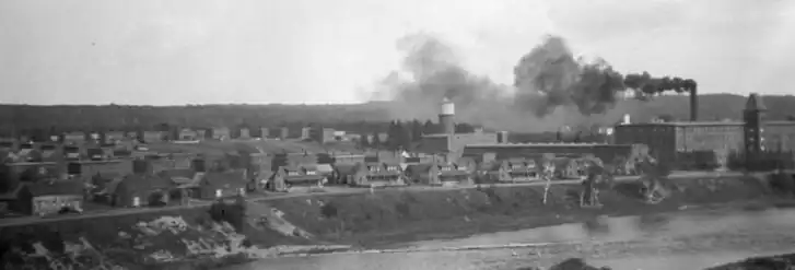 In the foreground is a river, with its far bank populated with many two-storey duplexes at left, and a large four-storey building to the right, behind which is a tall chimney releasing a black smoke billowing to the left.