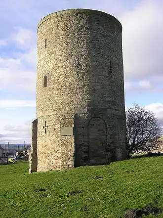 A three-story stone brick tower stands atop a grassy hill. The first story has a cross-shaped arrow slit, and the upper stories have small lancet windows.