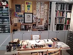 A desk covered with sketches, books, and art supplies, with walls behind displaying fashion drawings, photographs, and portraits