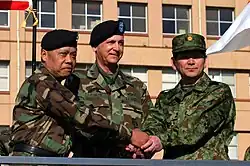 Two American generals wearing battle dress uniforms, and one Japanese general wearing a camouflage field uniform, all shaking hands.