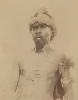 Portrait photograph of a shirtless Aboriginal man with a fur hat and a painted torso