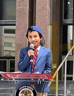 Photo of young boy with short black hair speaking at podium in front of building. He is wearing a business suit with blue suit jacket, necktie with alternating diagonal red and blue stripes, and a blue baseball cap turned backwards.