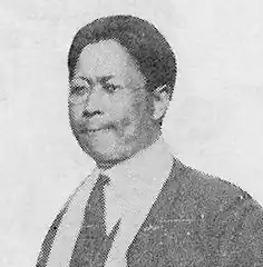 Black-and-white newspaper scan of a man in a suit and tie, wearing glasses. He has short black hair and has a slightly unpleased expression.