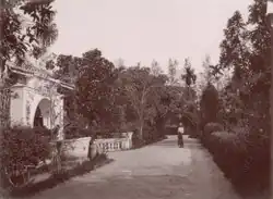 Path in a garden surrounded by trees and bushes. A side path on the left leads through a decorative arch in Chinese style. A man in Malay dress stands in the middle of the path.