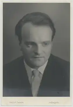 Black and white, three-quarter-profile, press shot of a clean-shaven man with brown hair brushed back, and a serious expression.