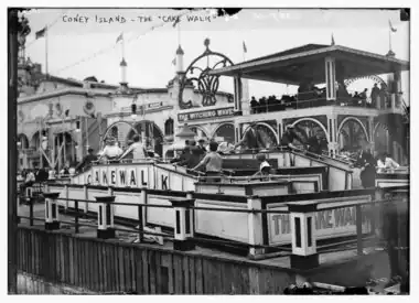 Old black and white photograph showing a fenced rectangle. In the rectangle, there are two aisles with ramps up into and down out from. About 8 people walk on the aisle closer to the viewer with their backs turned to the viewer. About 9 people walk in the opposite direction on the other rail. People are men, women and kids. All hold to the railing around the aisles. The ride is labelled "THE CAKE WALK" on one side and "C A K E W A L K" on another. Behind, a two-story ride labelled "THE WITCHING WAVE" and another probably labelled "SINKING OF THE MAINE". On the top left the photograph is labelled "CONEY ISLAND - THE "CAKE WALK"".