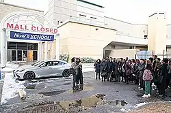 A group of people stand outside a vacant shopping mall. Above the doors, two signs on an awning read "Mall Closed" and "Now a School".
