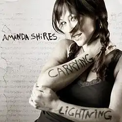 black and white half-length photo of Amanda Shires smiling as she displays words written on each arm, text "carrying" on one arm and "lightning" on the other