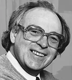 Black and white studio publicity portrait of a smiling, clean-shaven, bespectacled man with longish, straight hair with side parting and receding hairline