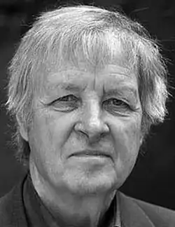 Black and white Press shot of an unsmiling, clean-shaven man, with longish white hair and fringe (bang).