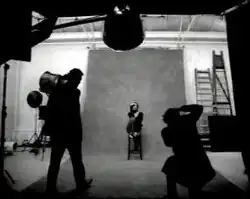 Lewis sits in a chair. She wears a sweater and jeans. In front of her one man is standing and another is taking photographs.