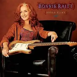 photo of Bonnie Raitt in orange print top and orange jacket with guitar seated in black leather armchair