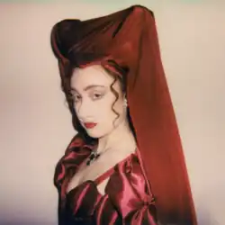 A three-quarter profile of Roan wearing a red ruffled dress, a red hood squared at the top, and a prosthetic pig nose.