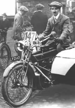 Collier sitting at the controls of his motorbike and sidecar at the start of the 1913 International Six Day Trial motorcycle endurance event. The sidecar with a passenger is to the right, mostly off-image