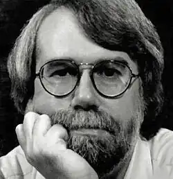Black and white publicity headshot of an unsmiling, bespectacled man with shoulder-length hair parted to one side, a trimmed beard, and full moustache. He is looking directly to camera, and resting his chin on his hand.