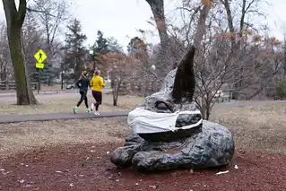 A bronze sculpture of a rabbit lying on its side on a bed of red mulch. Its ears stand tall and a cloth mask covers its nose and mouth. Behind it are a brown lawn, a pedestrian path upon which two joggers run, a parkway, and deciduous trees just starting to leaf.