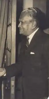Black and white photo of D. P. Atapattu wearing a suit and standing up, facing to the left and holding up his right arm.